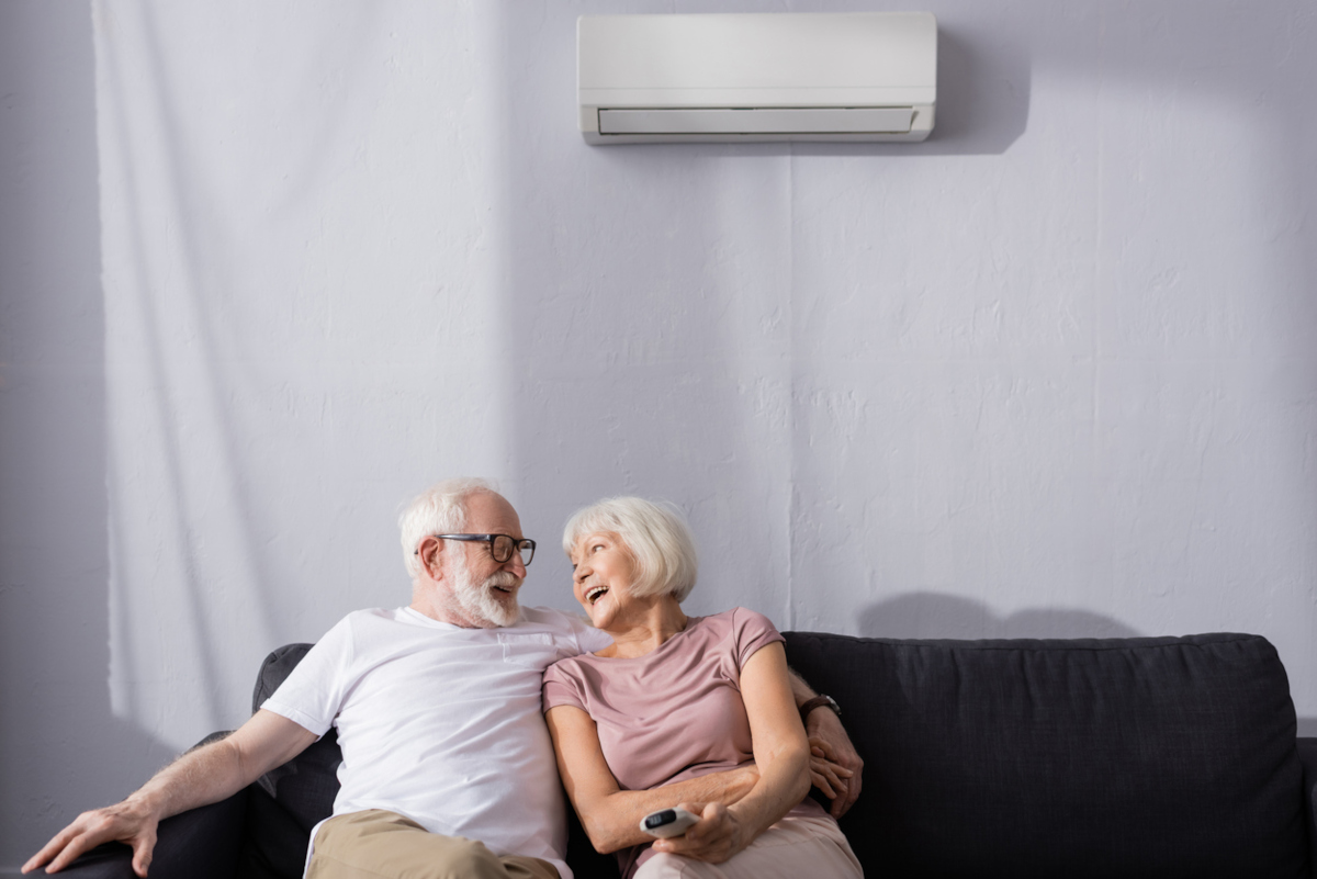 Ein älterer Mann mit Brille und weißem Haar sitzt am Sofa und umarmt dabei eine ältere Frau. Beide grinsen sich an. Oberhalb von ihnen is eine Klimaanlage zu sehen.