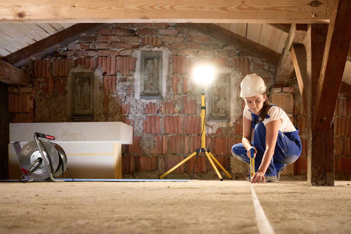 Eine Baurbeiterin misst im Dachboden die Flächen aus. Im Hintergund steht ein Scheinwerfer. Links neben ihr eine Kabeltrommel.