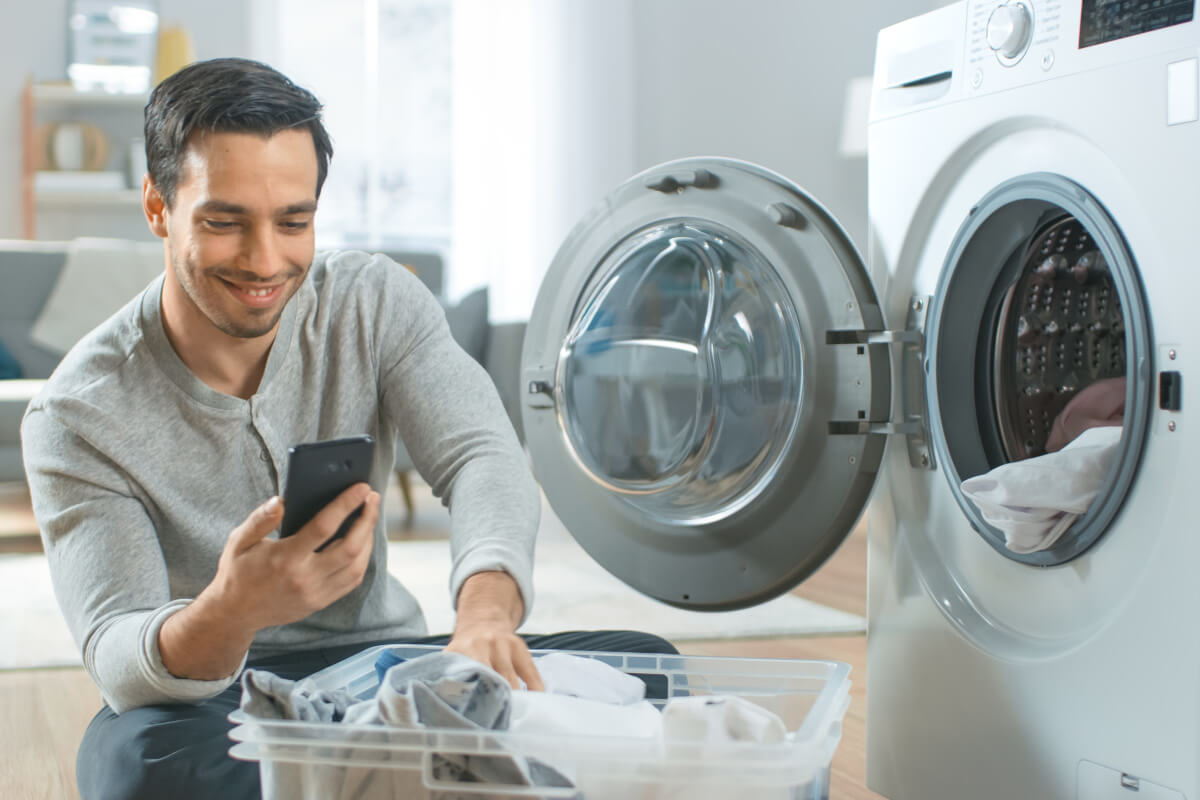 Hübsch lächelnde junge Mann in grauen Jeans und Mantel sitzt vor einer Waschmaschine und verwendet sein Smartphone. Er lädt Waschmaschine mit schmutziger Wäsche. Helles und geräumiges Wohnzimmer mit modernem Interieur.