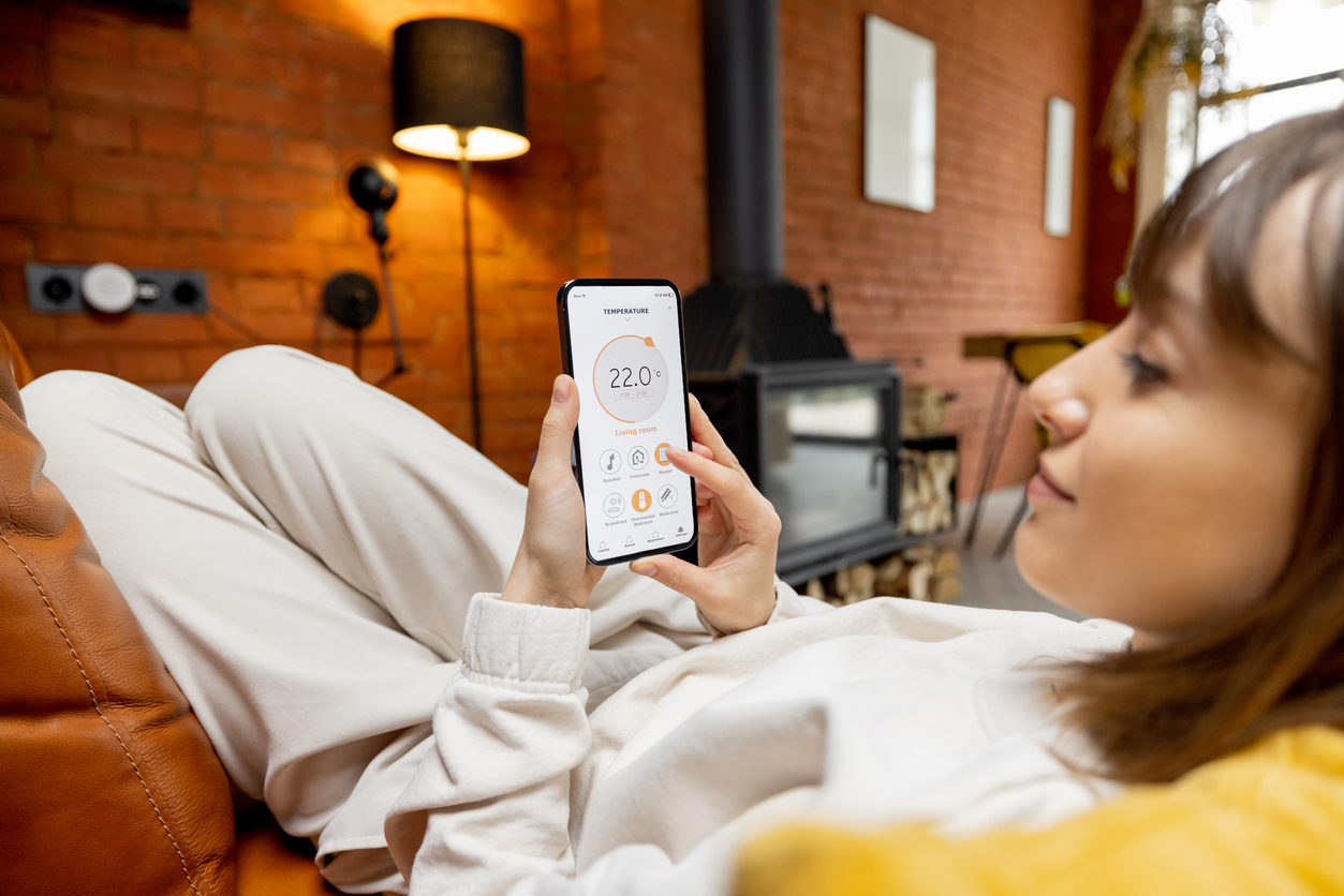 Eine Frau mit braunen Haaren liegt gemütlich auf der Couch und steuert mittels einer App am SMarthphone ihre Heizung.